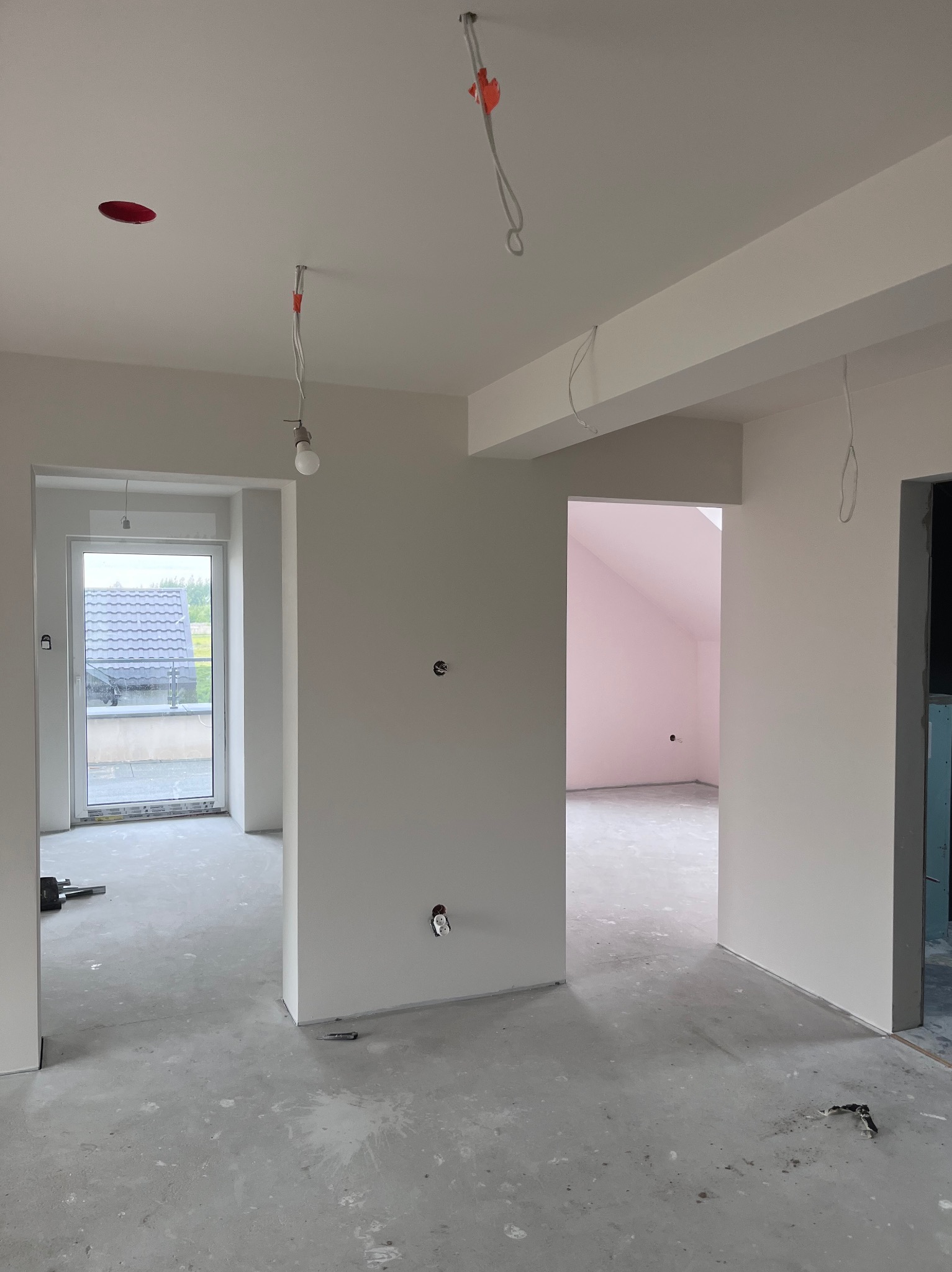 Wnętrze w trakcie prac wykończeniowych: świeżo pomalowane ściany na biało, surowa podłoga, widoczne instalacje elektryczne. W tle okno i różowe pomieszczenie.