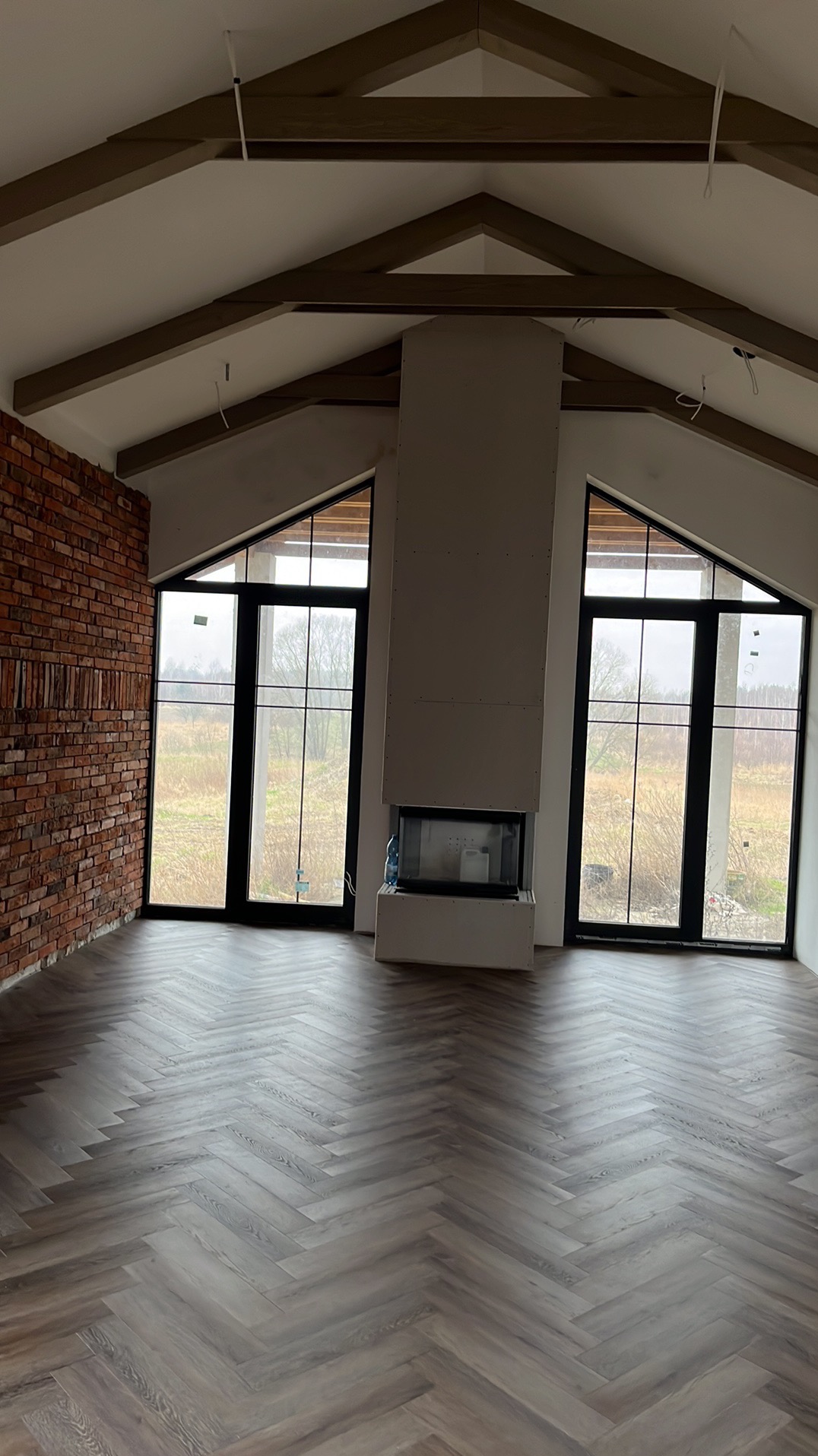 Jasne wnętrze z parkietem ułożonym w jodełkę, ceglaną ścianą i kominkiem obudowanym płytami gipsowymi. Duże okna z czarnymi ramami wpuszczają naturalne światło.