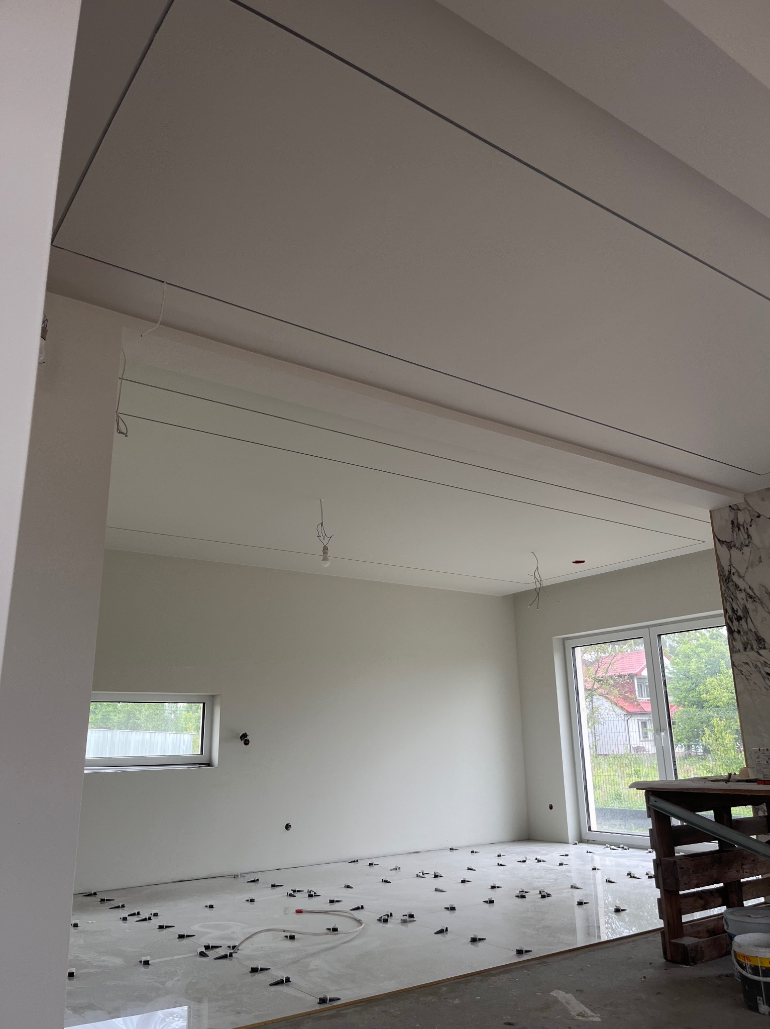 Jasne wnętrze w trakcie prac wykończeniowych: ułożone płytki podłogowe z elementami poziomującymi, podwieszony sufit z widocznymi przewodami. Okno i drzwi balkonowe.