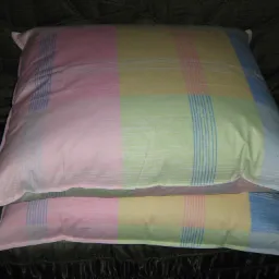 Dwa ręcznie pikowane kołdry ułożone jedna na drugiej, w pastelowych kolorach z wzorem w kratę i delikatnymi pionowymi paskami, leżące na ciemnym, teksturowanym tle.