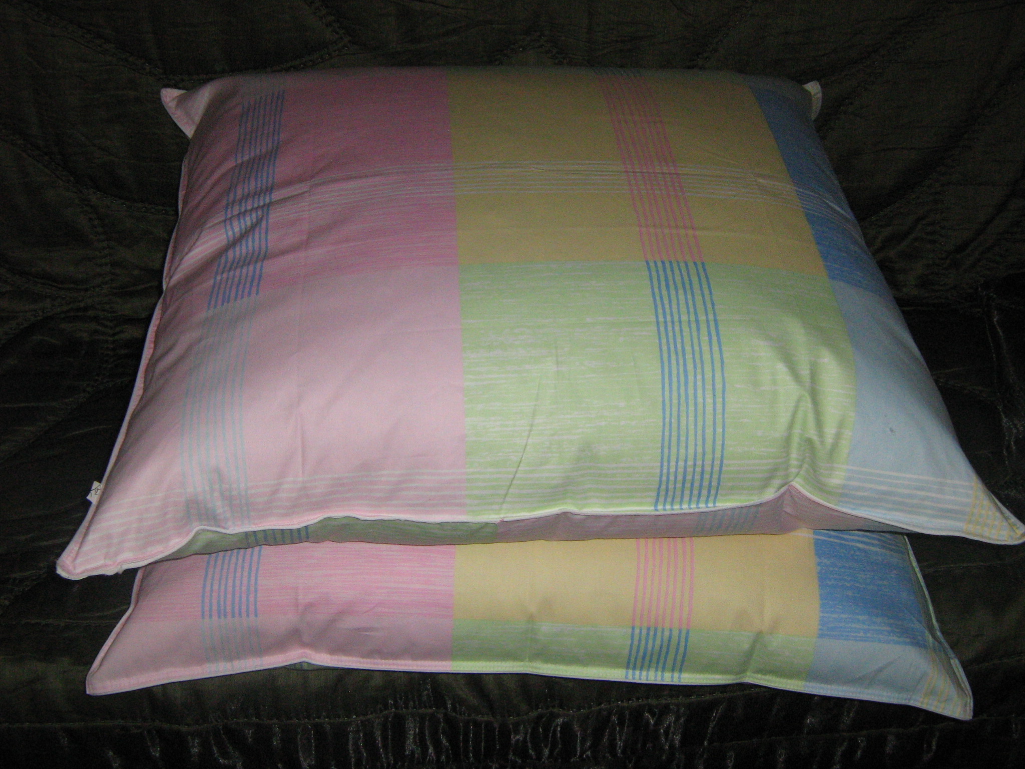 Dwa ręcznie pikowane kołdry ułożone jedna na drugiej, w pastelowych kolorach z wzorem w kratę i delikatnymi pionowymi paskami, leżące na ciemnym, teksturowanym tle.