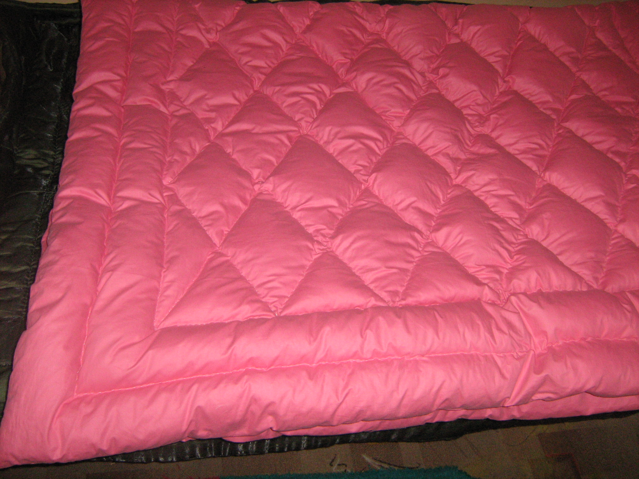 Ręcznie pikowana kołdra w odcieniu różu, z geometrycznym wzorem szwów tworzącym trójkąty, leżąca na ciemnej, błyszczącej tkaninie.