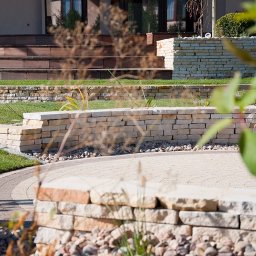 Ogrodomania Łukasz Nadgrodkiewicz - Ogród z kamiennymi murkami oporowymi, ścieżką z kostki brukowej w odcieniach beżu i brązu oraz schodami z drewnianych desek prowadzącymi do domu.