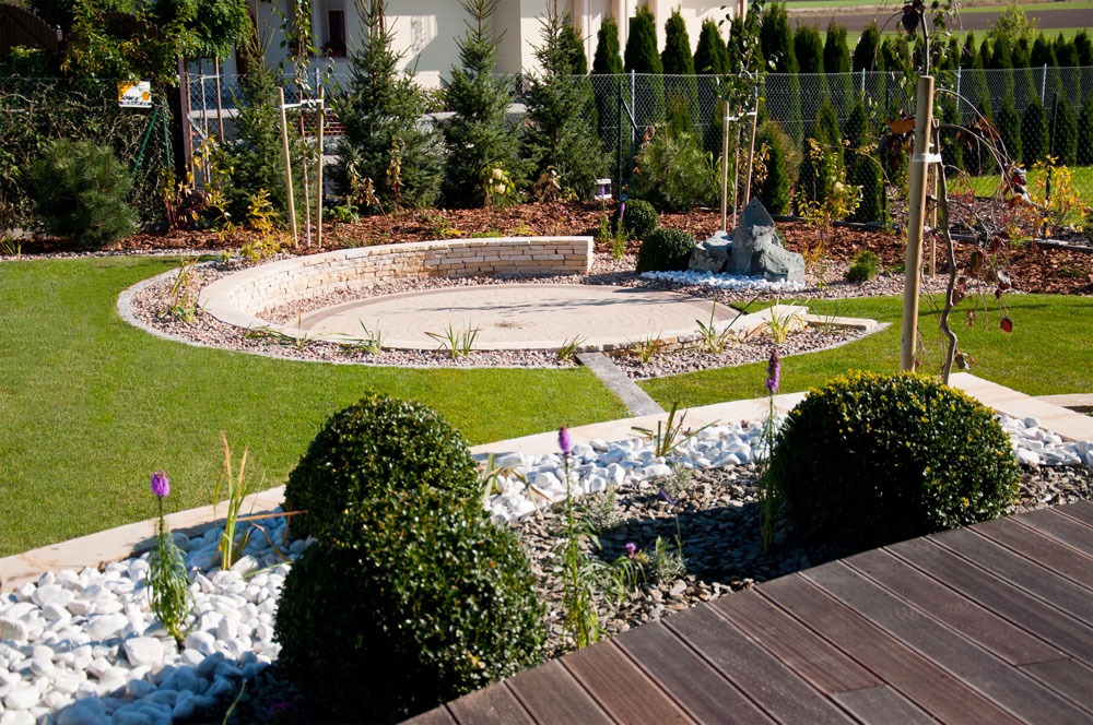 Starannie zaprojektowany ogród z okrągłym placem wyłożonym drobnym żwirem, otoczonym niskim murem z piaskowca i obrzeżem z kostki, trawnik, rabaty z białym grysem, kuliste krzewy i drewniany taras...