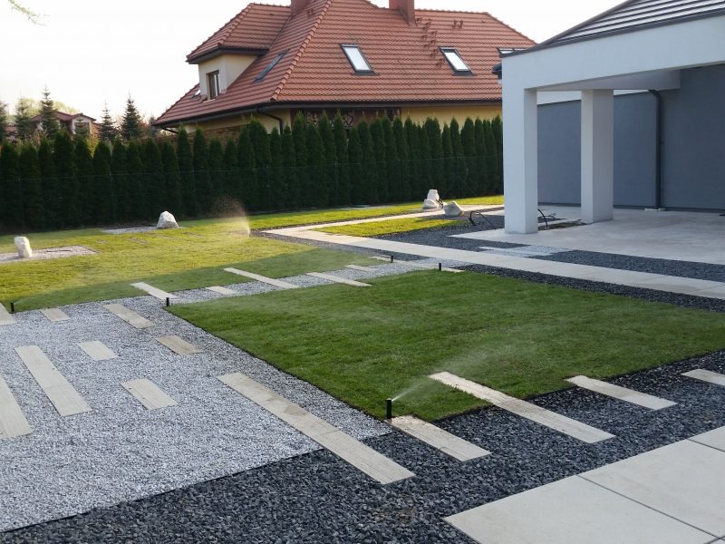 Nowoczesny ogród z automatycznym systemem nawadniania w trakcie pracy, widoczne zraszacze, ścieżki z jasnego i ciemnego grysu, płyty chodnikowe, trawnik, tuje i dom w tle.