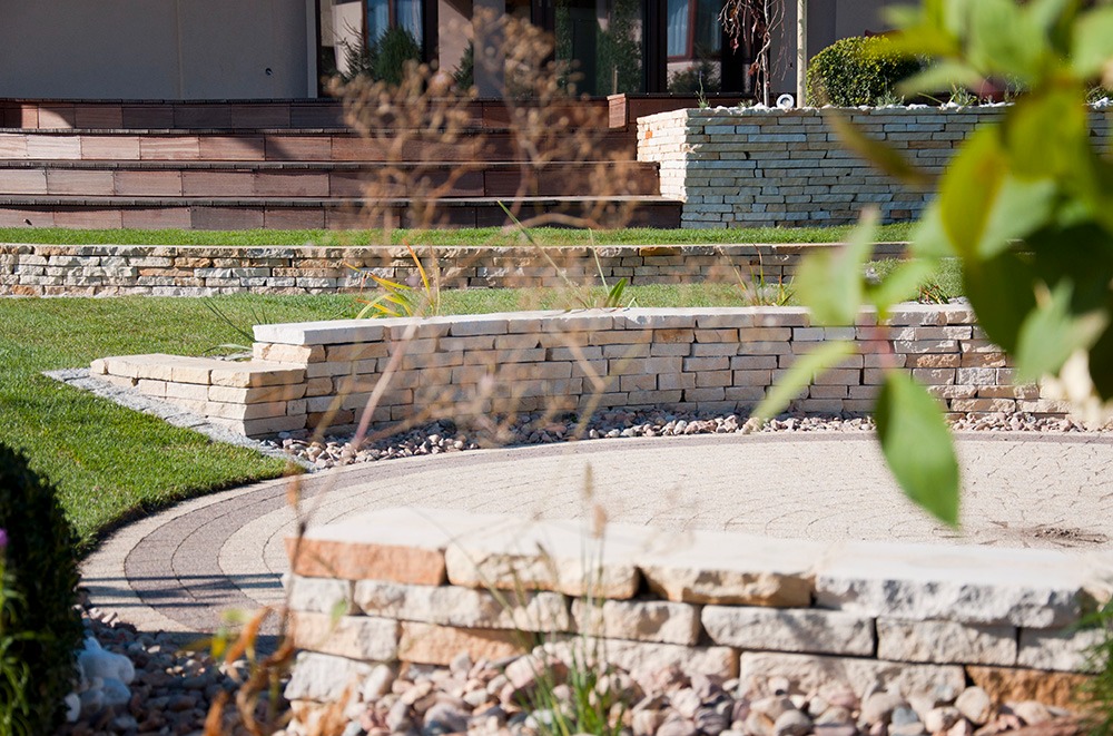 Ogród z kamiennymi murkami oporowymi, ścieżką z kostki brukowej w odcieniach beżu i brązu oraz schodami z drewnianych desek prowadzącymi do domu.