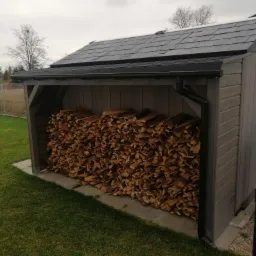 Drewniana wiata na drewno opałowe z dachem pokrytym gontem bitumicznym, z rynną, ustawiona na betonowej płycie, na tle trawnika i drzew.