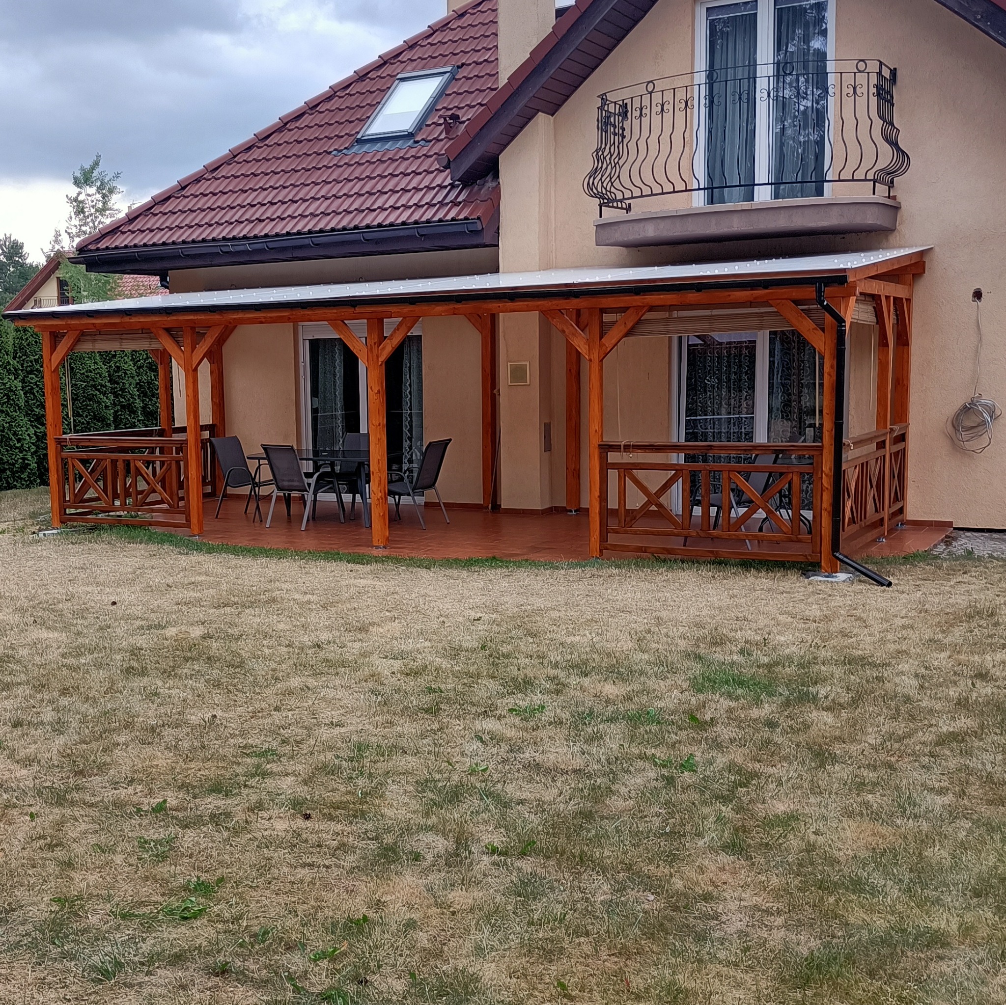 Zadaszony taras z drewnianą konstrukcją, wyposażony w meble ogrodowe, przylegający do domu z balkonem i czerwoną dachówką, otoczony trawnikiem.