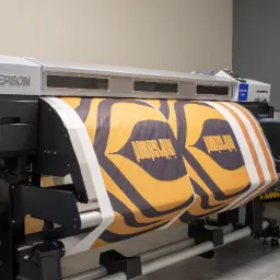 Drukarka wielkoformatowa Epson drukuje wzór w odcieniach pomarańczy i czerni na białym materiale, widoczny komputer z oprogramowaniem drukarki w tle.