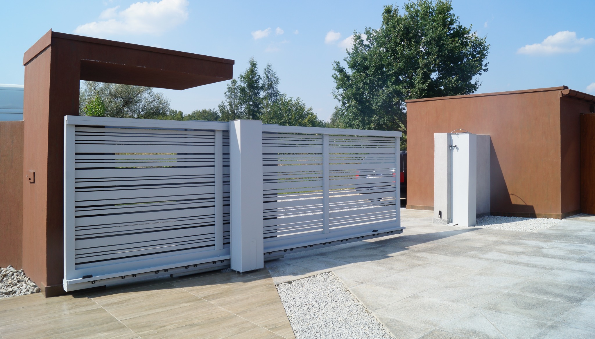 Nowoczesna, przesuwna brama w kolorze szarym z poziomymi szczelinami, zamontowana na posesji z brązowymi elementami architektonicznymi i jasną nawierzchnią z płytek.