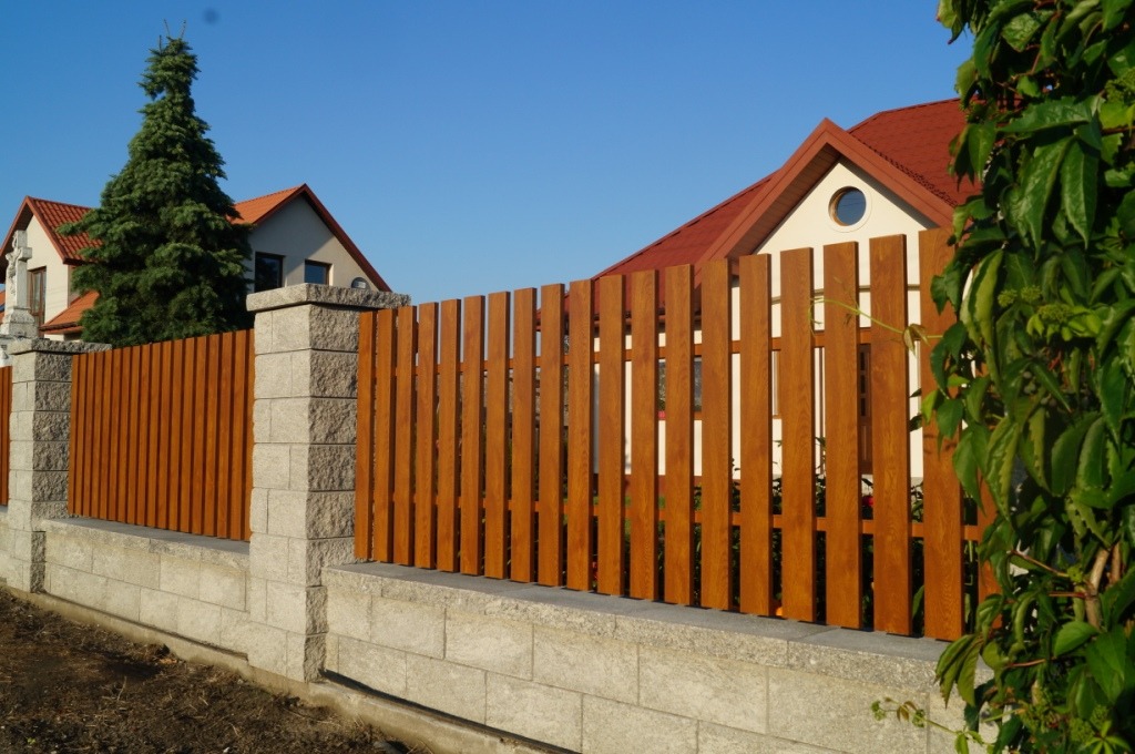 Nowoczesne, drewniane ogrodzenie panelowe z betonową podmurówką i kamiennymi słupkami, na tle domów jednorodzinnych i błękitnego nieba.