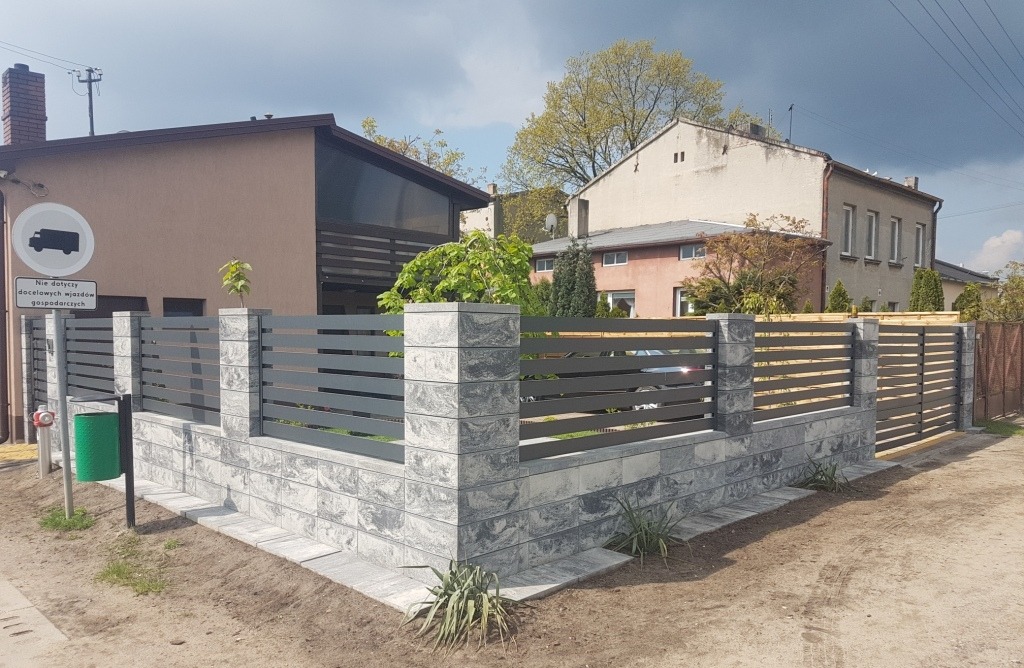 Nowoczesne ogrodzenie z poziomych, szarych paneli metalowych i murku z szarych bloczków betonowych, z widocznym znakiem drogowym i koszem na śmieci w tle zabudowy mieszkalnej.