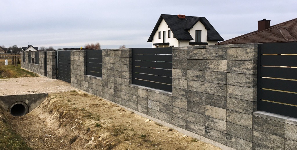 Nowoczesne ogrodzenie z szarego kamienia łupanego z poziomymi, czarnymi przęsłami, widok z zewnątrz na tle domów jednorodzinnych i pochmurnego nieba.