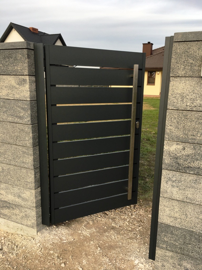 Nowoczesna, otwarta brama wjazdowa z poziomymi panelami, zamontowana między dwoma słupkami z szarego betonu architektonicznego, widok na posesję z domem w tle.