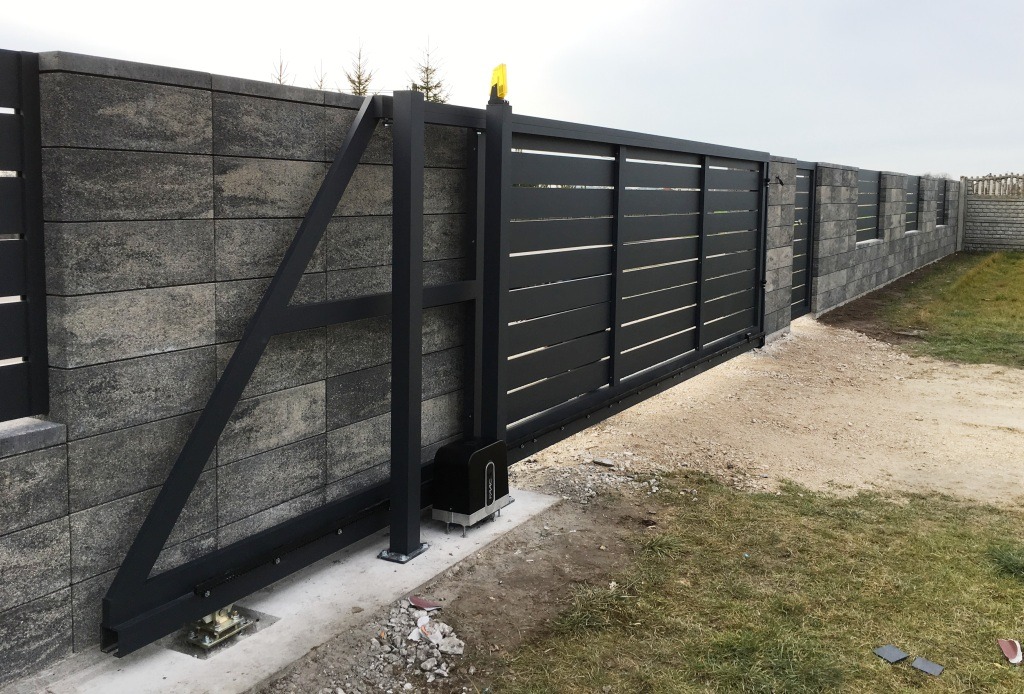 Nowoczesna, przesuwna brama metalowa z automatyką, zamontowana przy ogrodzeniu z bloczków betonowych w szarym odcieniu. Widoczny mechanizm napędowy bramy oraz lampka ostrzegawcza.
