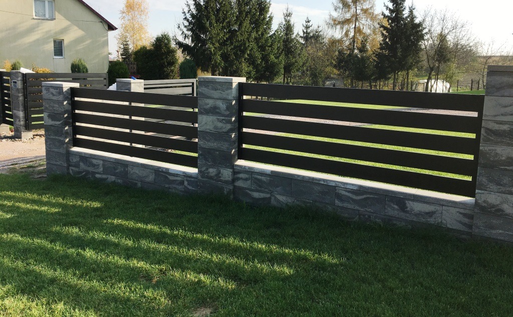 Nowoczesne, poziome ogrodzenie metalowe w kolorze antracytowym, z kamiennymi słupkami i podmurówką w odcieniach szarości, otoczone zielonym trawnikiem i zabudową jednorodzinną.