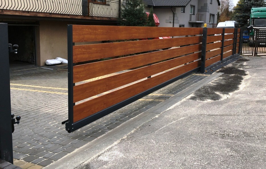 Przesuwna brama wjazdowa z poziomych desek w kolorze drewna, osadzona na czarnej, metalowej ramie, widoczna na tle kostki brukowej i budynku z garażem.