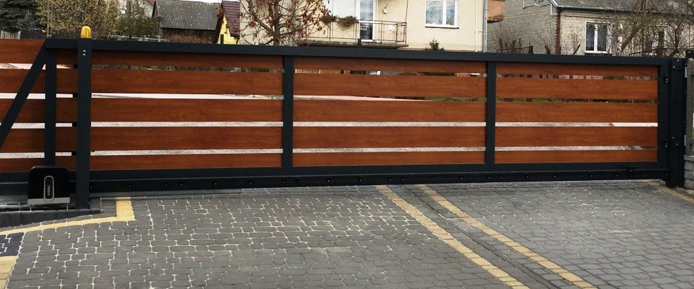 Przesuwna brama z poziomych desek w odcieniach brązu, osadzona w czarnej, metalowej ramie, z widocznym napędem elektrycznym, na posesji wyłożonej kostką brukową.