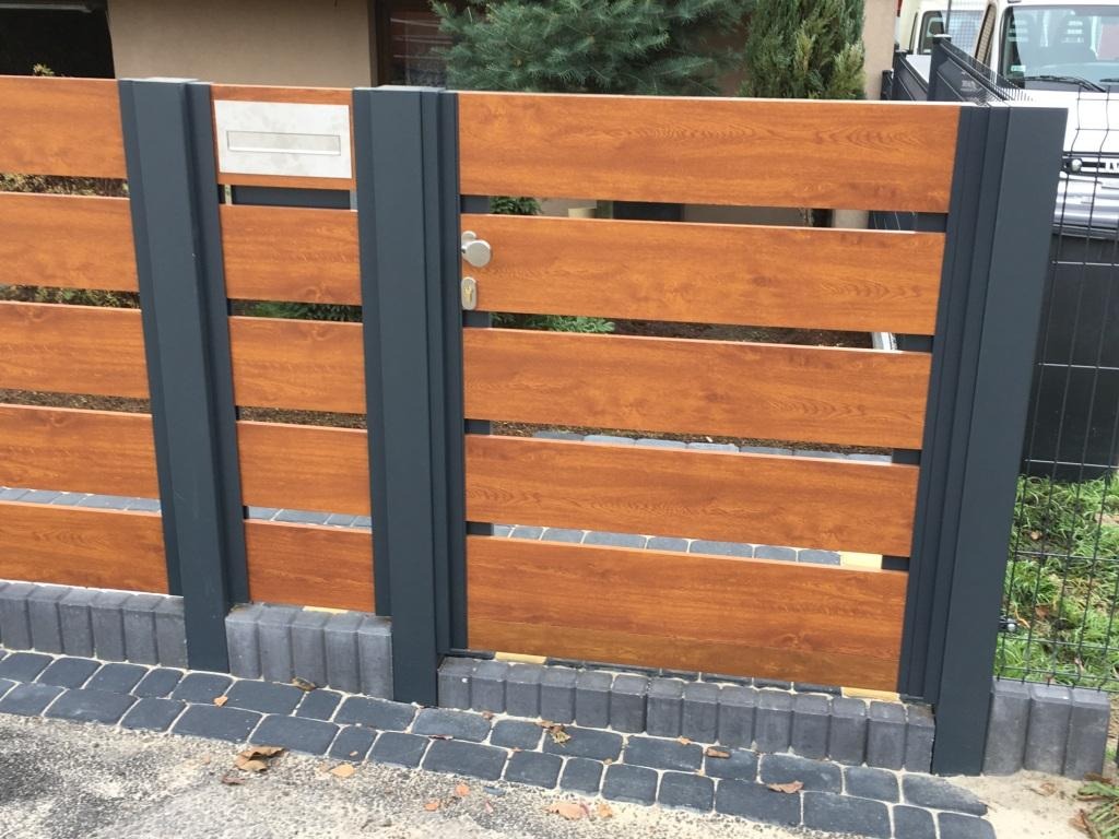 Nowoczesne ogrodzenie panelowe z drewnopodobnymi przęsłami w kolorze ciepłego brązu i grafitowymi słupkami, zamontowane na podmurówce z kostki brukowej, widoczna skrzynka na listy i furtka.