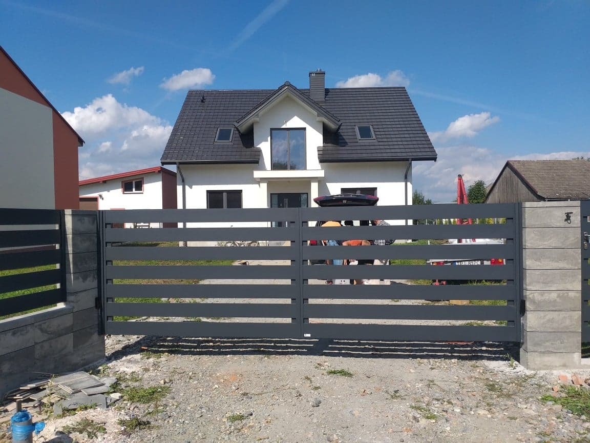 Nowoczesna, pozioma brama przesuwna z szarego metalu na tle białego domu z ciemną dachówką i błękitnego nieba z chmurami.