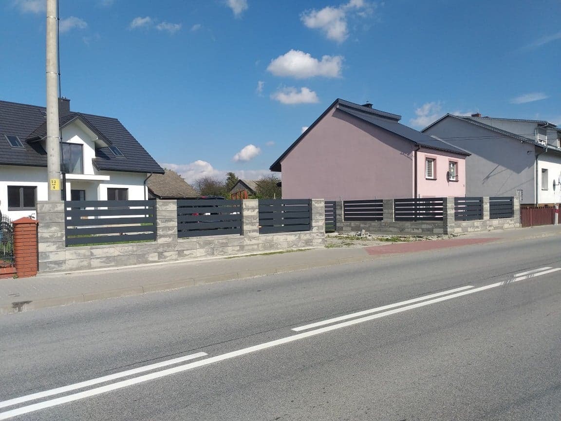 Nowoczesne ogrodzenie panelowe z szarego metalu na betonowej podmurówce z elementami imitującymi kamień, oddzielające posesje od ulicy w słoneczny dzień.