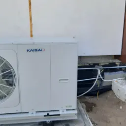 Zewnętrzna jednostka pompy ciepła Kaisai zamontowana przy ścianie budynku z otwartymi drzwiami wejściowymi, widoczne przewody i betonowe bloczki podtrzymujące instalację.