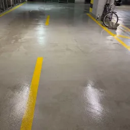 Błyszcząca, szara posadzka betonowa z żółtymi liniami w garażu podziemnym, obok zaparkowany rower i torba.