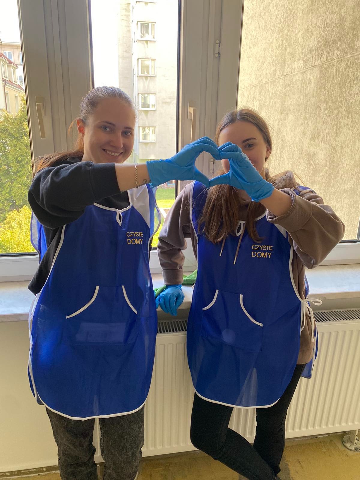 Dwie uśmiechnięte kobiety w niebieskich fartuchach z napisem 'Czyste Domy' i niebieskich rękawiczkach formują dłońmi kształt serca na tle okna z widokiem na budynek.