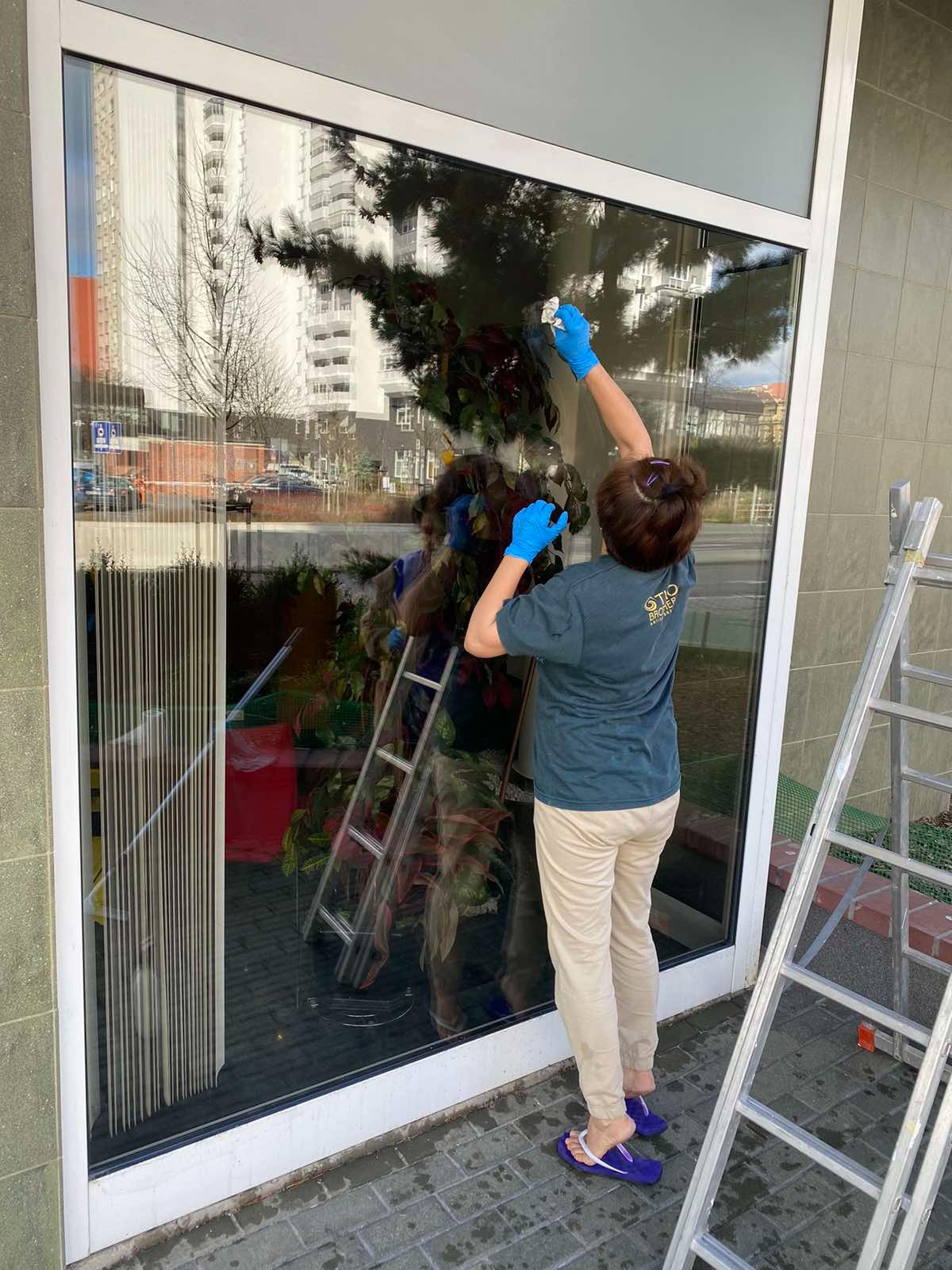 Osoba w niebieskich rękawicach, z widocznym logo firmy na koszulce, myje okno na parterze budynku, obok stoi rozłożona drabina.