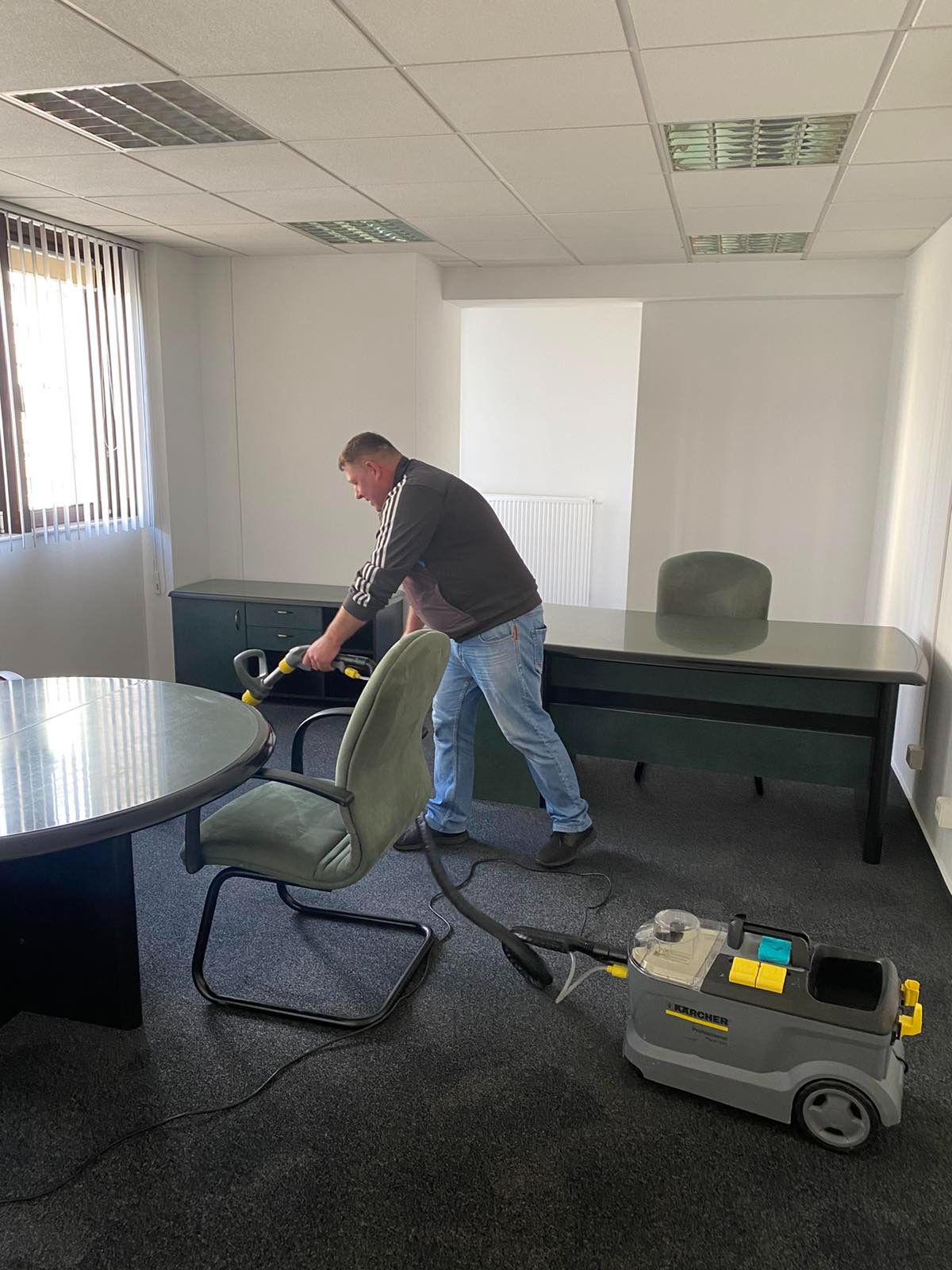 Mężczyzna w dresie czyści ciemną wykładzinę w biurze za pomocą profesjonalnego odkurzacza piorącego Karcher, widoczne biurko, krzesła i okno z żaluzjami.