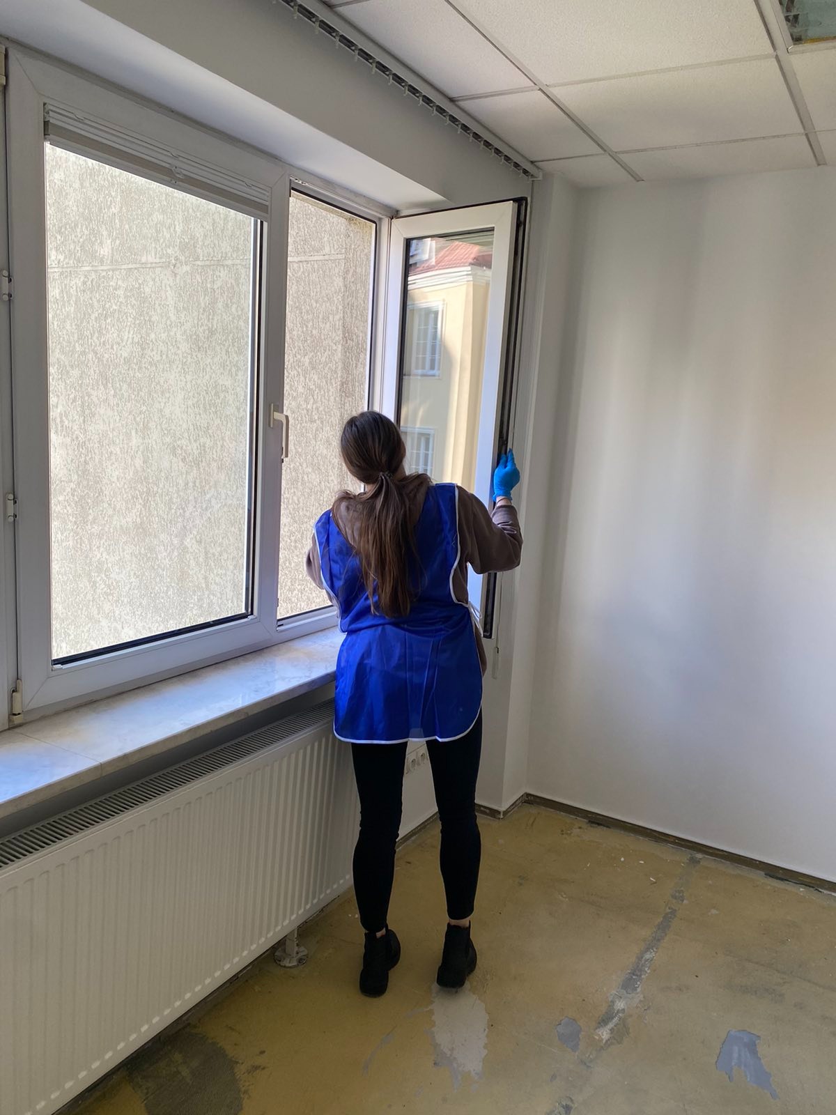 Kobieta w niebieskim fartuchu i rękawiczkach myje otwarte okno w jasnym pomieszczeniu z grzejnikiem i częściowo zniszczoną podłogą.