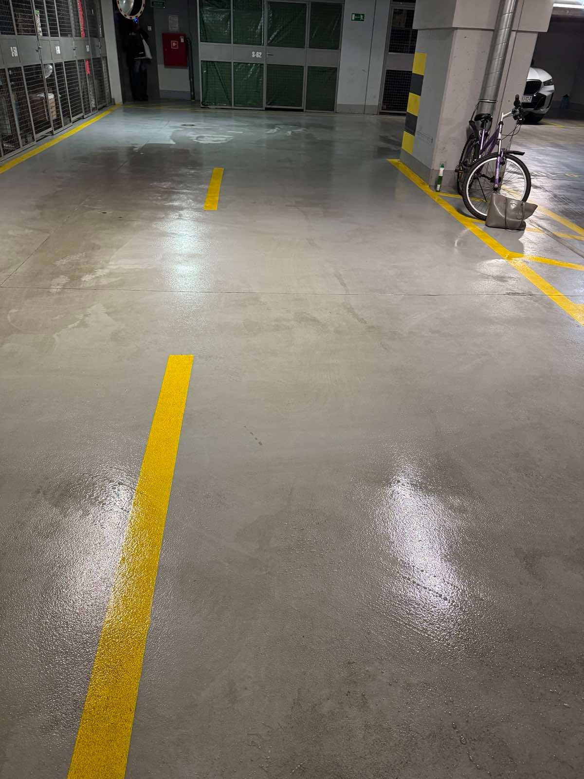 Błyszcząca, szara posadzka betonowa z żółtymi liniami w garażu podziemnym, obok zaparkowany rower i torba.