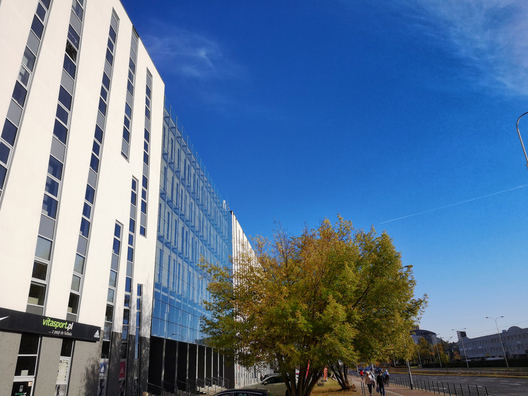 Nowoczesny budynek biurowy z przeszkloną fasadą i białymi elementami, widziany z poziomu ulicy w słoneczny dzień, z drzewem na pierwszym planie i widocznym sklepem sportowym na parterze.