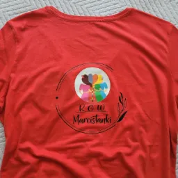 Czerwona koszulka z nadrukiem przedstawiającym stylizowane postacie kobiet z warkoczami w okręgu i napisem 'KGW Marcistanki' poniżej, leżąca na wzorzystej tkaninie.
