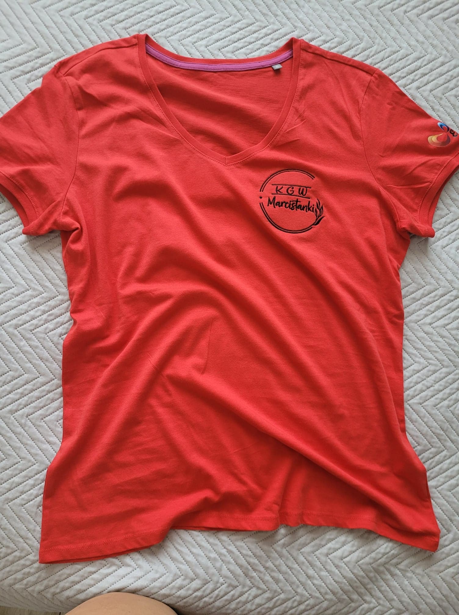 Czerwona koszulka z krótkim rękawem i dekoltem w serek, z czarnym nadrukiem 'KGW Marcistanki' w okręgu oraz kolorowym logo na rękawie, leżąca na białym, wzorzystym tle.
