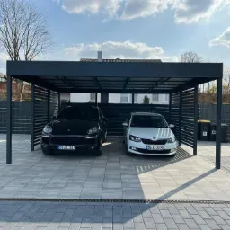 Nowoczesna, dwustanowiskowa wiata samochodowa z metalową konstrukcją i lamelowymi ścianami bocznymi, chroniąca czarny SUV i białe auto osobowe na posesji z kostki brukowej.