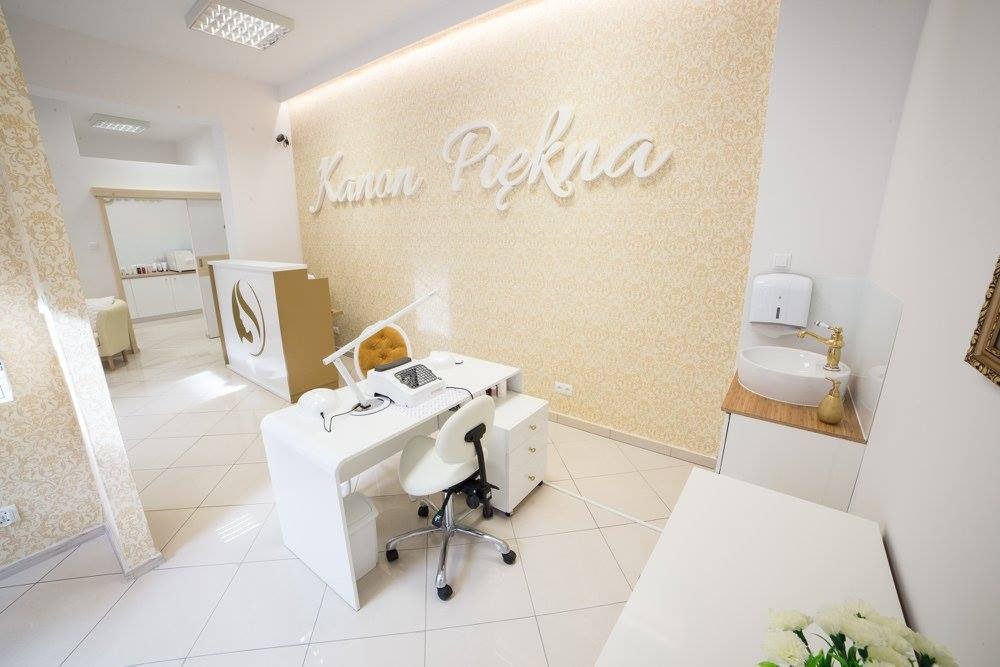 Jasne wnętrze salonu z białymi meblami, lampą do manicure i napisem 'Kanon Piękna' na ścianie w tle.
