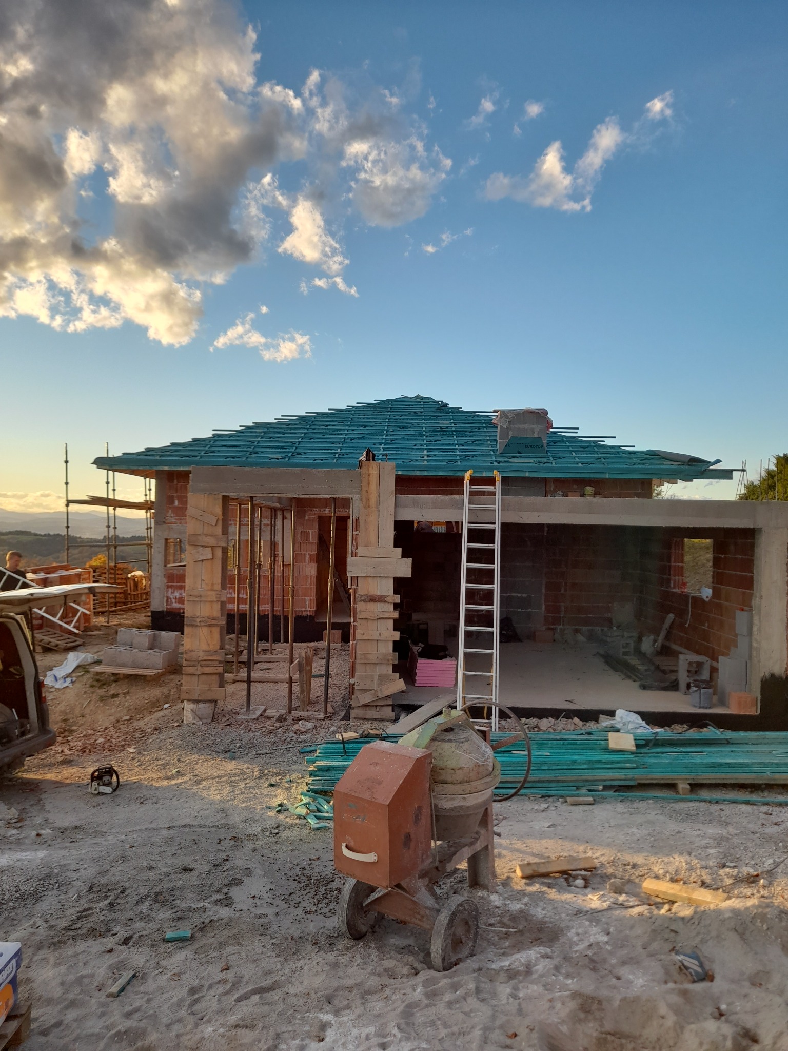 Surowy stan budowy domu z cegły z zielonym dachem w trakcie budowy, widoczna betoniarka i drabina, na tle błękitnego nieba z chmurami.