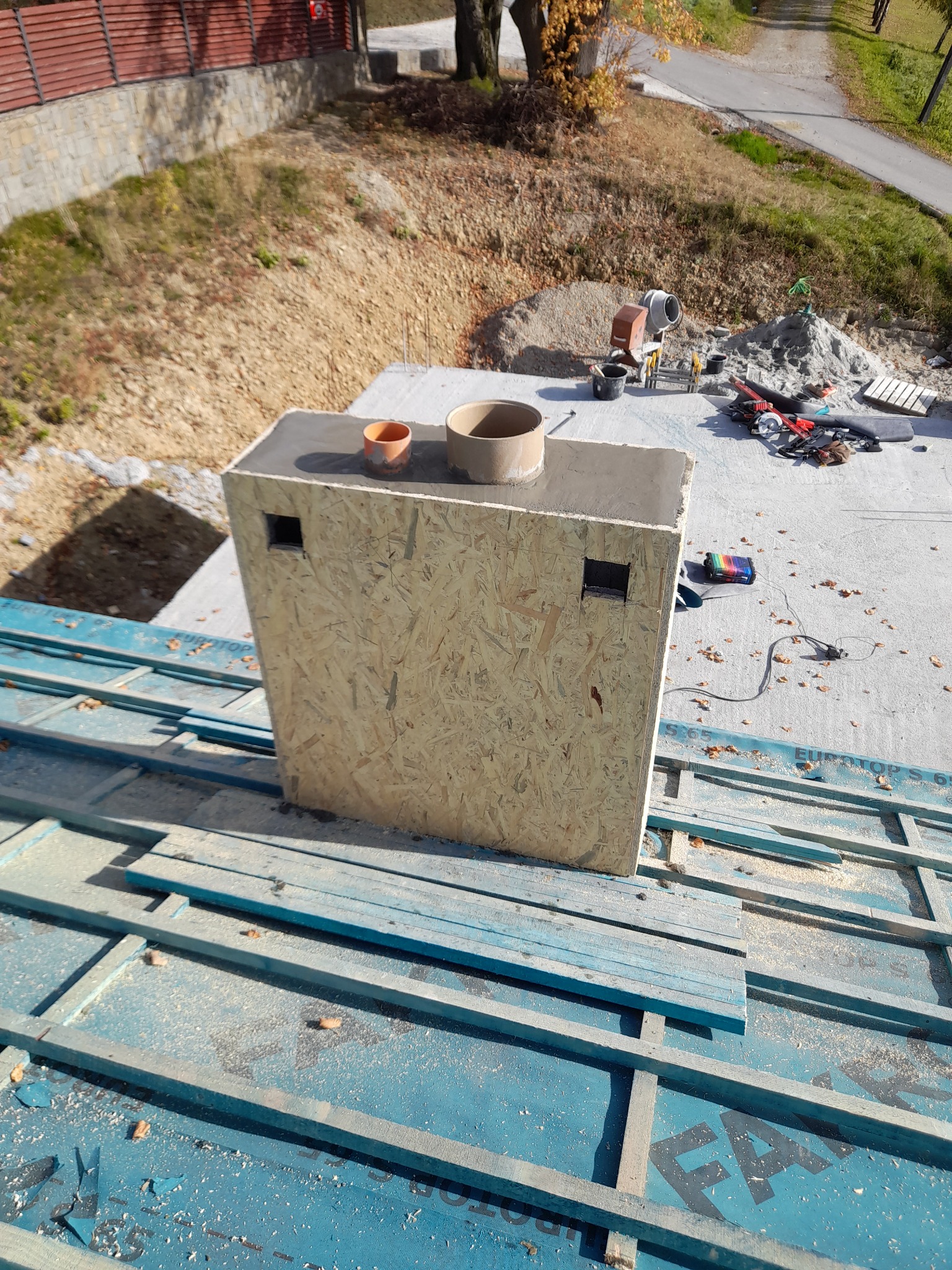 Budowa komina: drewniana forma z otworami na dachu z niebieską membraną, widoczny mikser do betonu i narzędzia w tle.