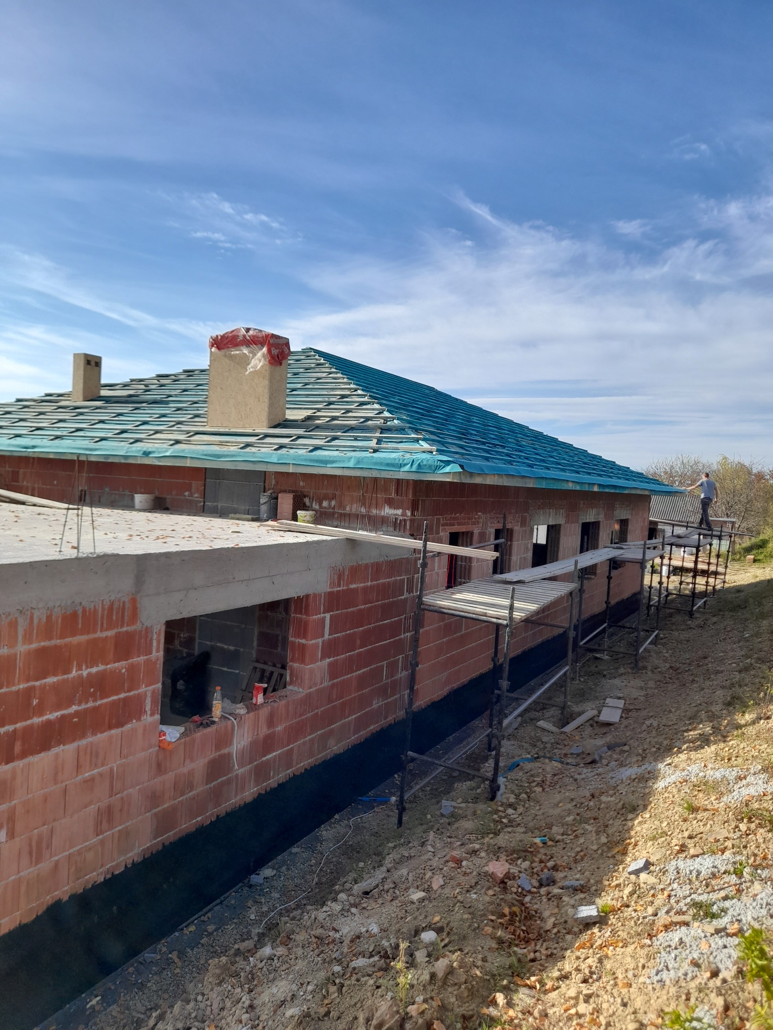 Surowy budynek z czerwonej cegły w trakcie budowy, z prowizorycznym dachem z niebieskiej folii, widoczne rusztowania i fragment krajobrazu z osobą na nich.
