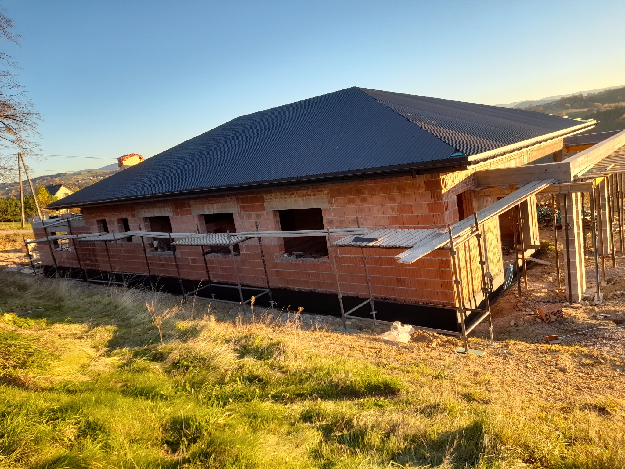 Surowy stan domu z czerwonej cegły z ciemnym dachem z blachy falistej, widoczne rusztowania i krajobraz w tle.