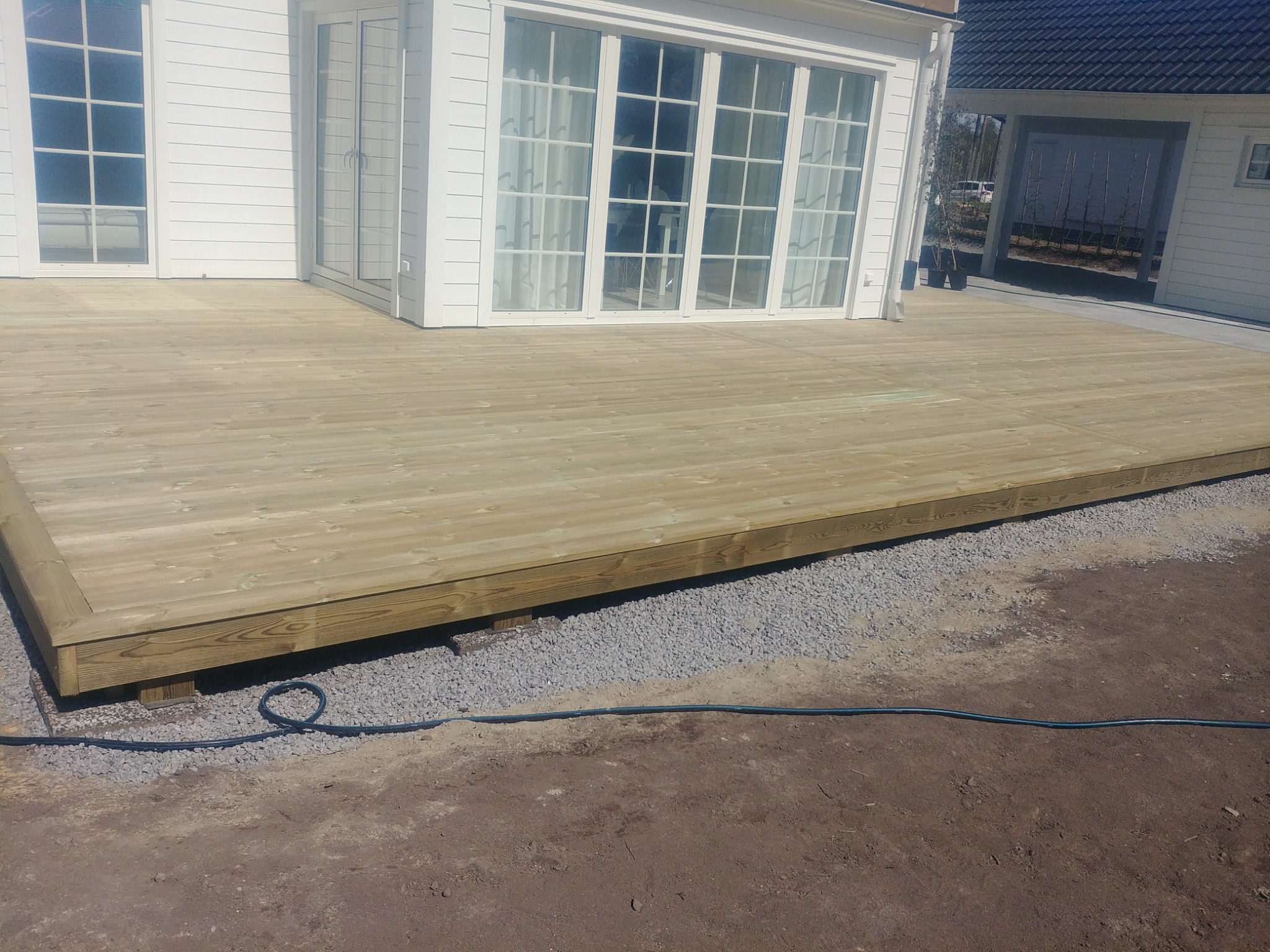 Nowo wybudowany taras z jasnego drewna, przylegający do białego domu z dużymi oknami, widoczne podsypka z kamieni i kabel na ziemi.