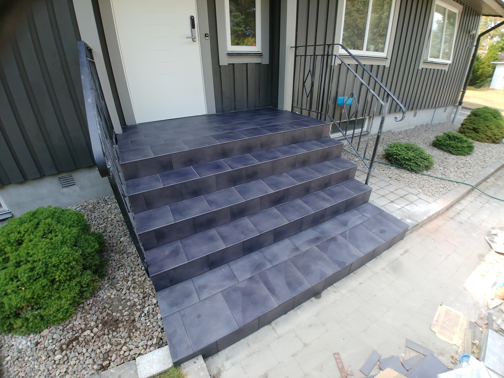 Zewnętrzne schody wejściowe wyłożone ciemnymi płytkami ceramicznymi z metalową barierką, widok na elewację domu z szarą boazerią i białymi oknami, obok ozdobne krzewy i kamyki.