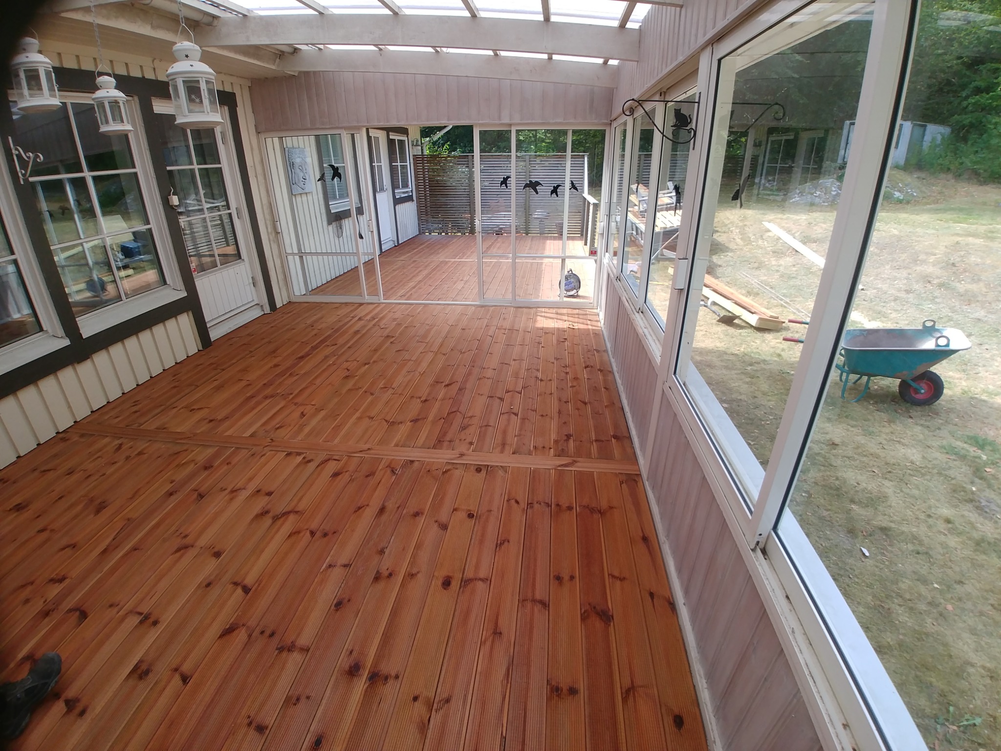 Zadaszony taras z drewnianą podłogą i przeszklonymi ścianami, widok na ogród z taczką w tle.