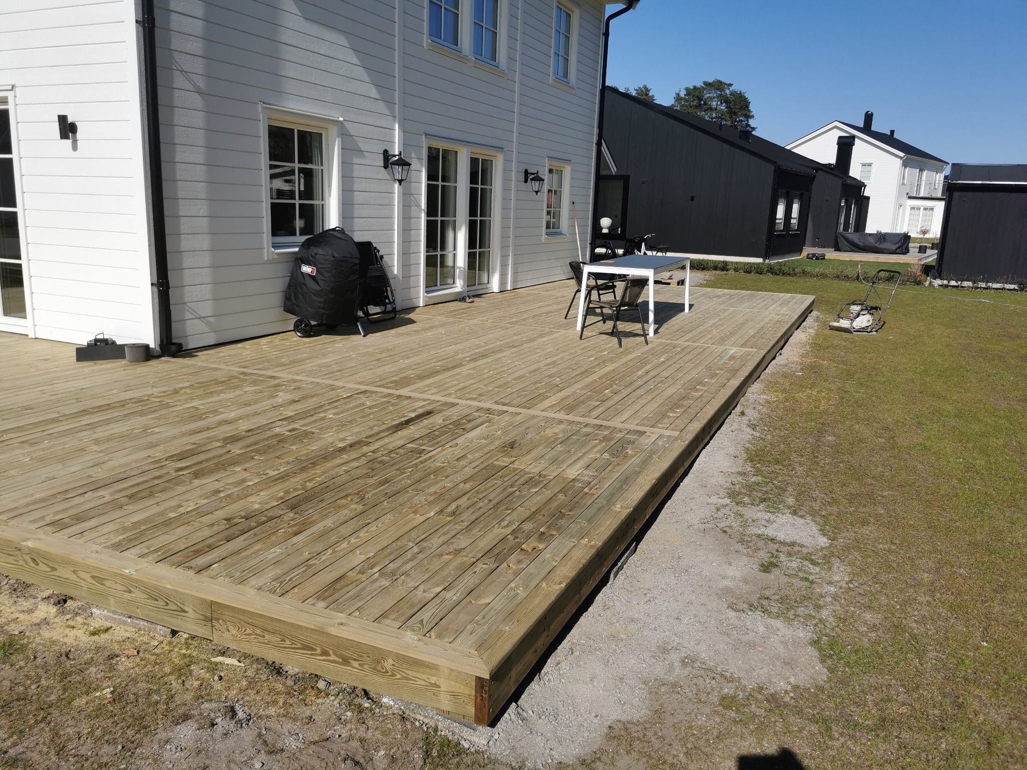 Nowo wybudowany taras z jasnego drewna przy białym domu z czarnymi akcentami, widoczny stół z krzesłami, grill w pokrowcu, trawnik i maszyna do aeracji gleby w tle.