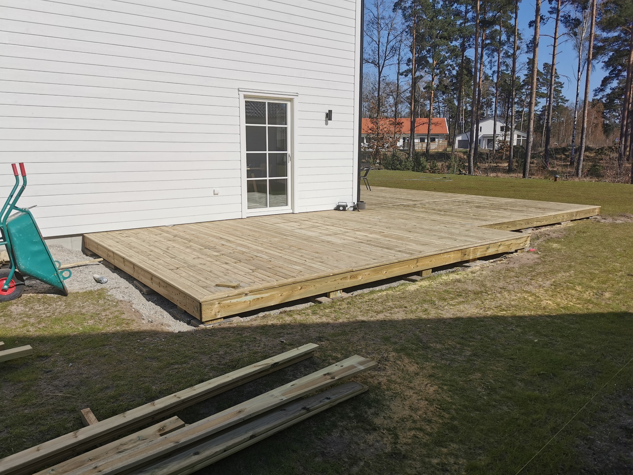 Drewniany taras w trakcie budowy przy białym domu, widoczna taczka i deski, drzewa w tle.