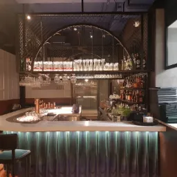 Profesjonalnie wyposażony bar z podświetlanym frontem, szklankami i butelkami na metalowej konstrukcji, zielone krzesła barowe w tle.