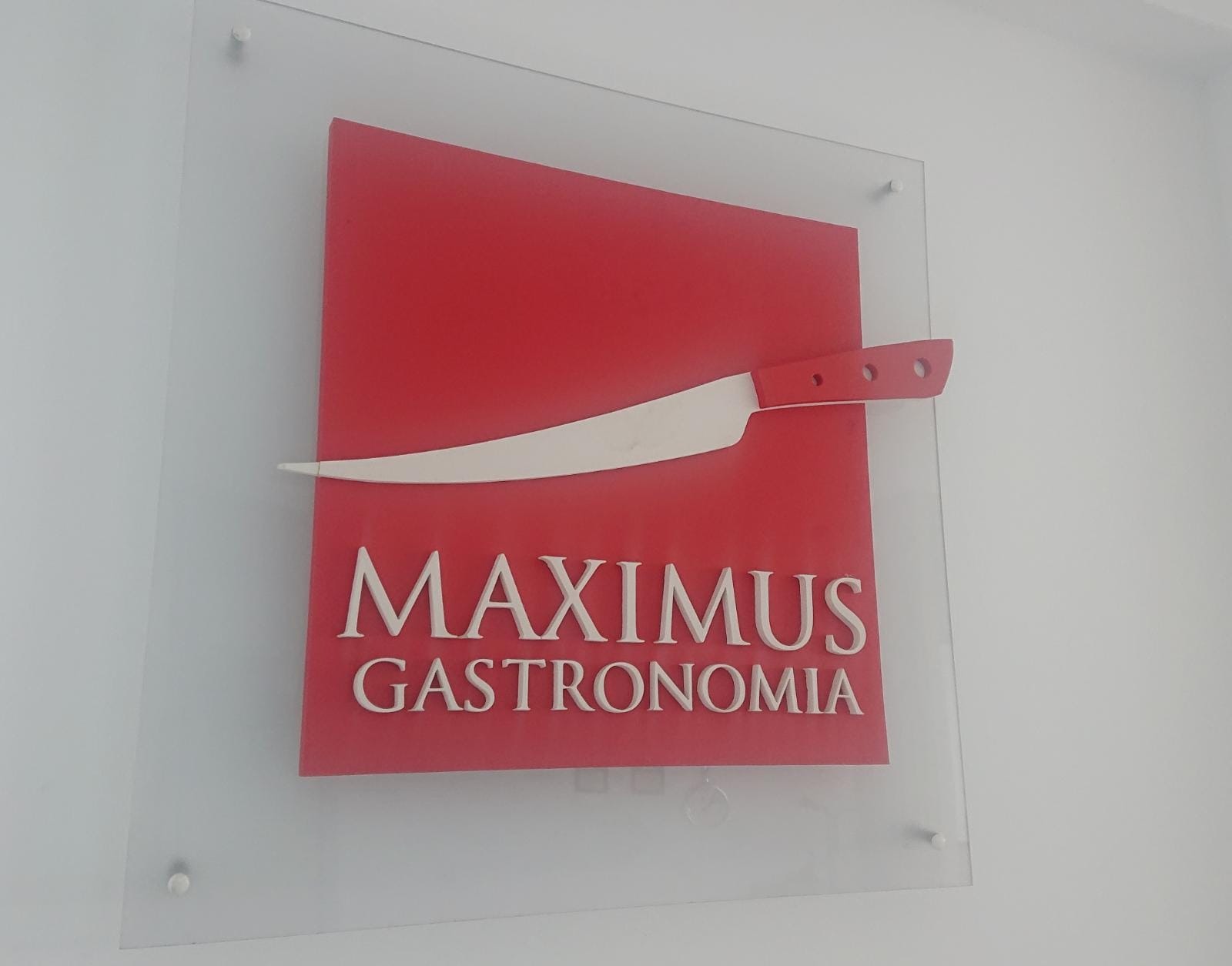 Logo firmy Maximus Gastronomia z czerwonym kwadratem, białym nożem i białym napisem, zamocowane na szklanej płycie na białej ścianie.