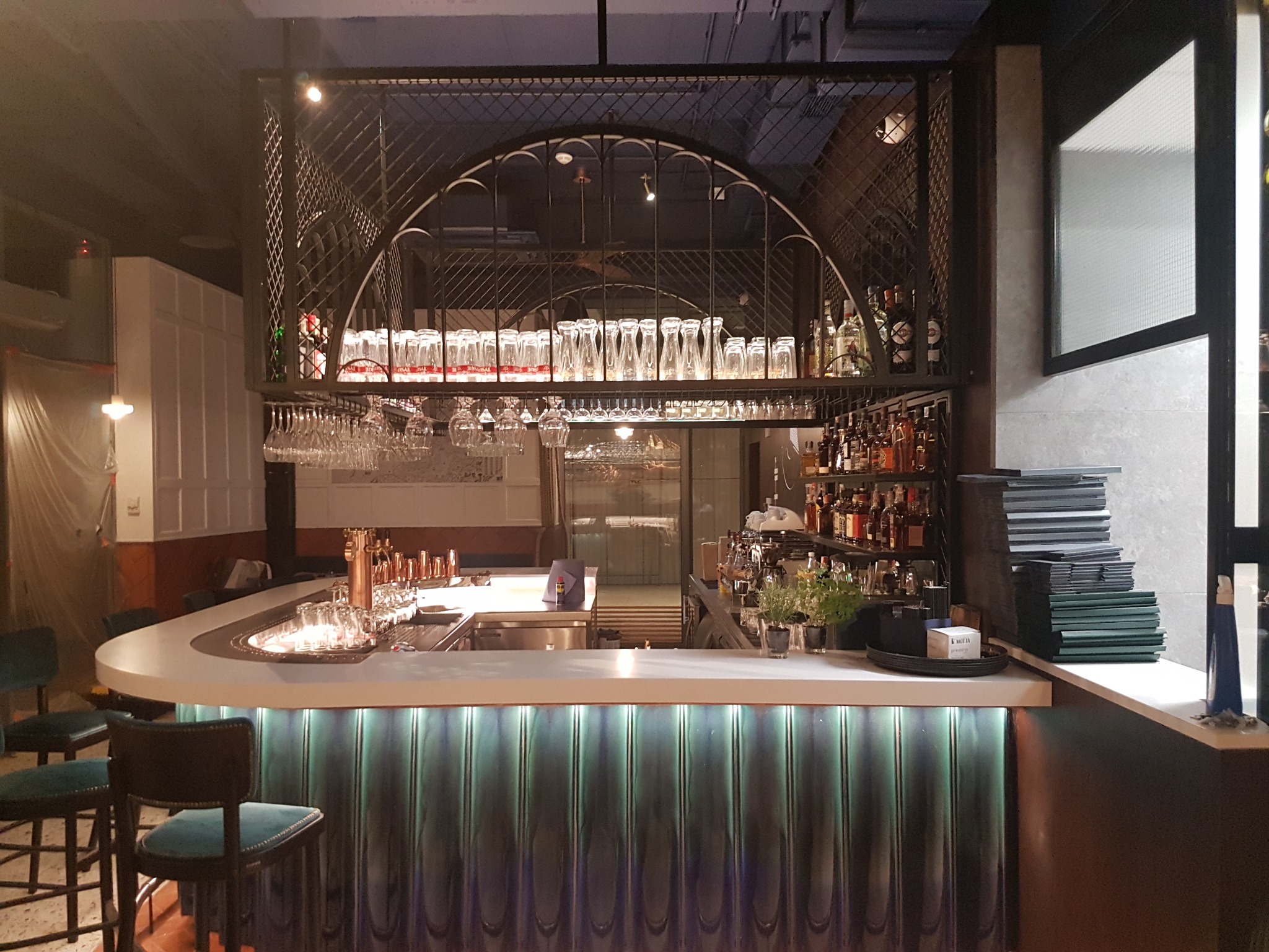 Profesjonalnie wyposażony bar z podświetlanym frontem, szklankami i butelkami na metalowej konstrukcji, zielone krzesła barowe w tle.