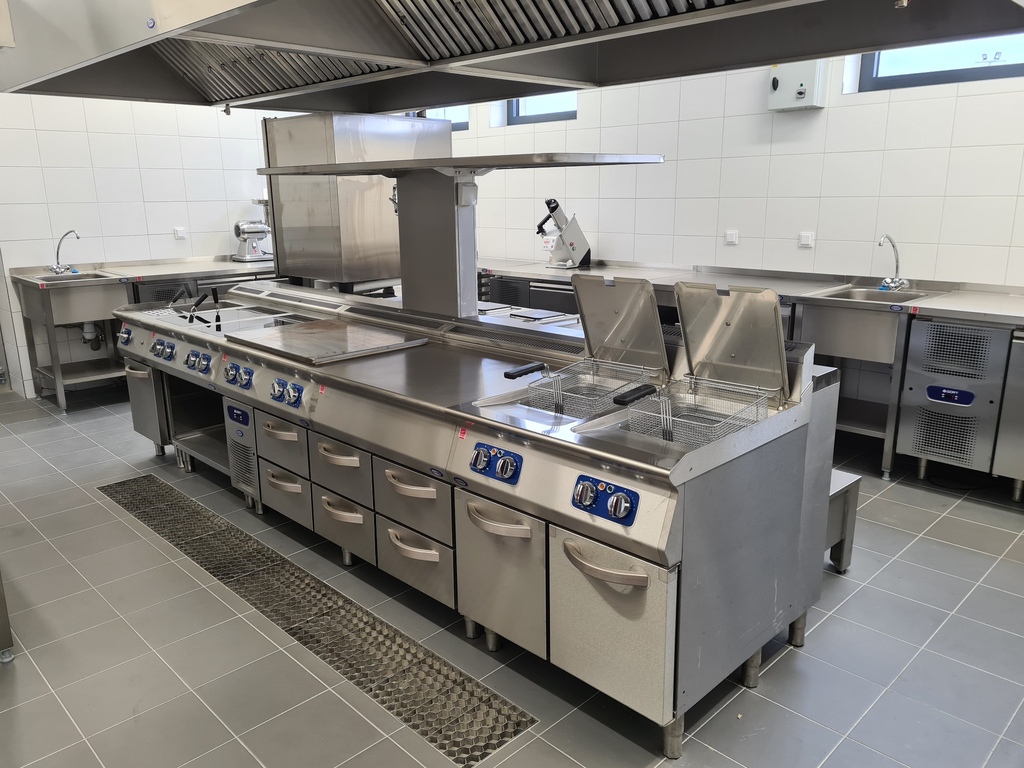 Profesjonalna, stalowa kuchnia gastronomiczna z frytownicami, płytą grzewczą i okapem w jasnym pomieszczeniu z białymi kafelkami, widoczne również zlewozmywaki i inne urządzenia.